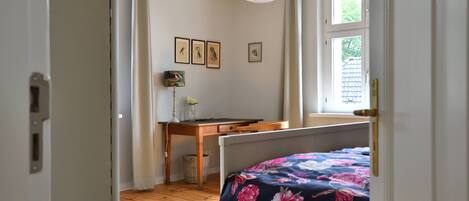 Une chambre avec un plancher en bois, un lit recouvert d’une couvre-lit à motifs floraux, un bureau avec une lampe et des tableaux encadrés accrochés au mur.