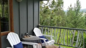 Une terrasse en bois avec deux chaises Adirondack blanches et une petite table, donnant sur une zone boisée.