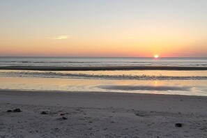 Uma praia ao pôr do sol, com mar calmo e céu claro.