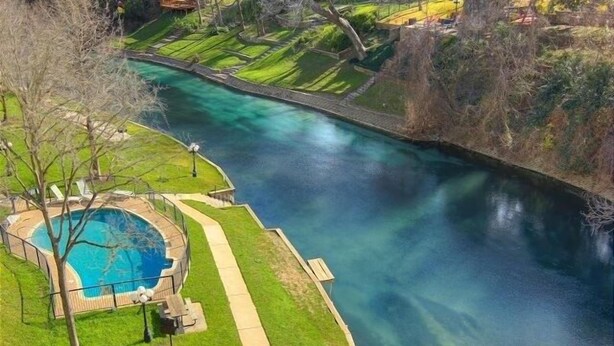 A swimming pool with a clear blue water feature, surrounded by a well-maintained lawn and a walking path. - Cowboy Corral Ic 112 2 Bedroom Condo by RedAwning (New Braunfels)