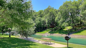 A picnic area with a table and benches by a calm river, surrounded by lush greenery. - Cowboy Corral Ic 112 2 Bedroom Condo by RedAwning (New Braunfels)