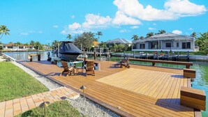 A wooden deck with lounge chairs and a grill, overlooking a marina with boats and a house in the background. - Southern Exposure Waterfront Home | Pool & Dock (Marco Island)