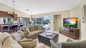 A spacious living room with a large sofa, a coffee table, and a television. There is a dining area with chairs and a table, and a kitchen in the background. - Southern Exposure Waterfront Home | Pool & Dock (Marco Island)