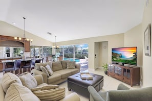 A spacious living room with a large sofa, a coffee table, and a television. There is a dining area with chairs and a table, and a kitchen in the background.