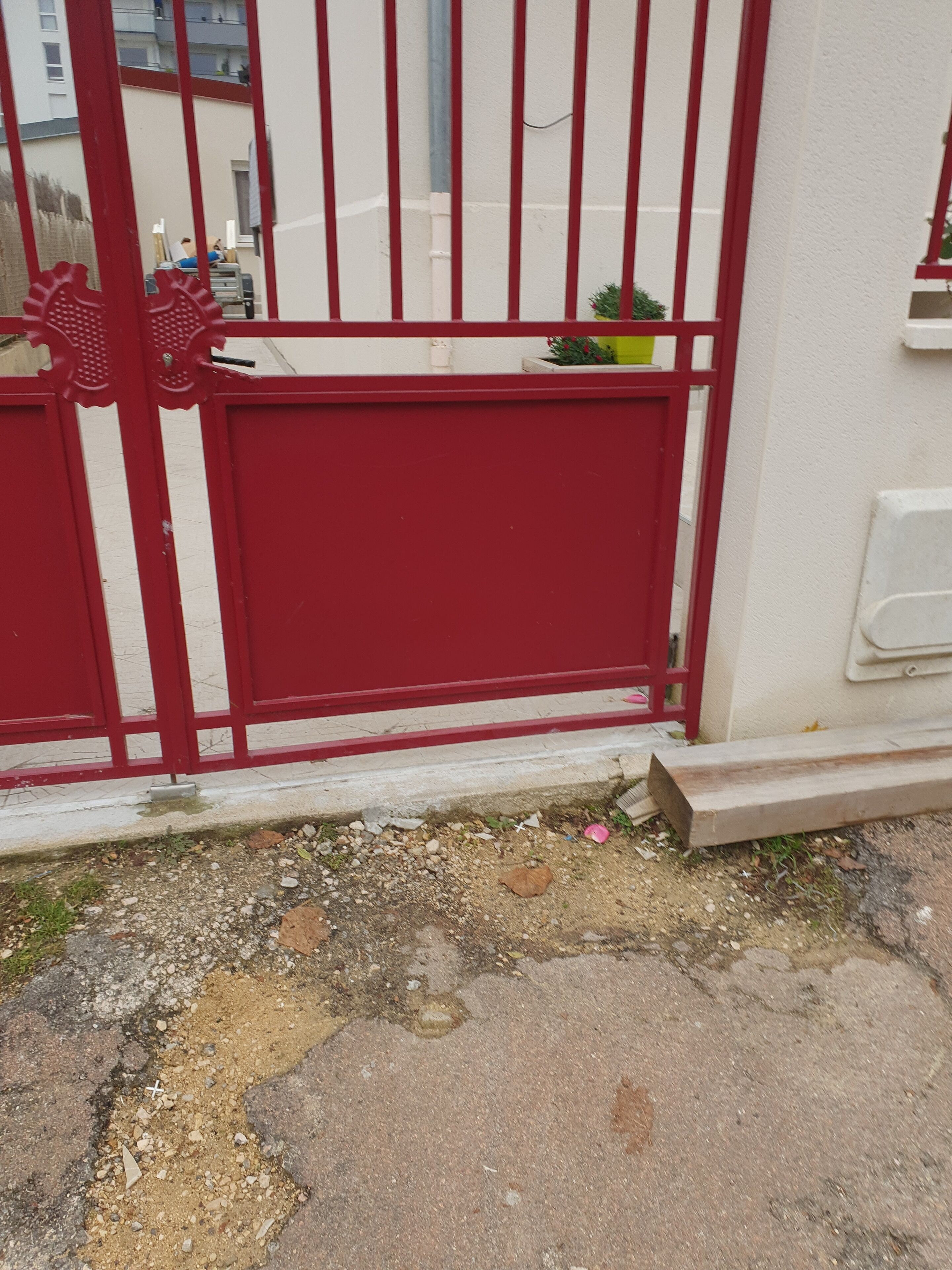 A red metal gate with a decorative top, a concrete base, and a small potted plant to the right.
