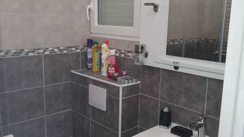 Baño con inodoro, lavamanos y ducha. Hay artículos de aseo en el lavamanos y una ventana sobre el espejo.