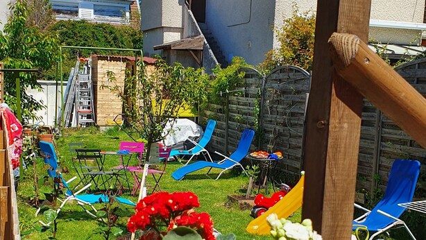 Un parterre surélevé en bois, agrémenté de fleurs roses et blanches, une pergola en bois et des chaises de jardin bleues dans un jardin.