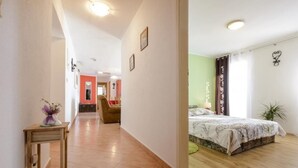 A hotel room with a bed, a small table with a vase, a chair, and a view of a hallway with a red door. - Stone House Kuna (Konavle)