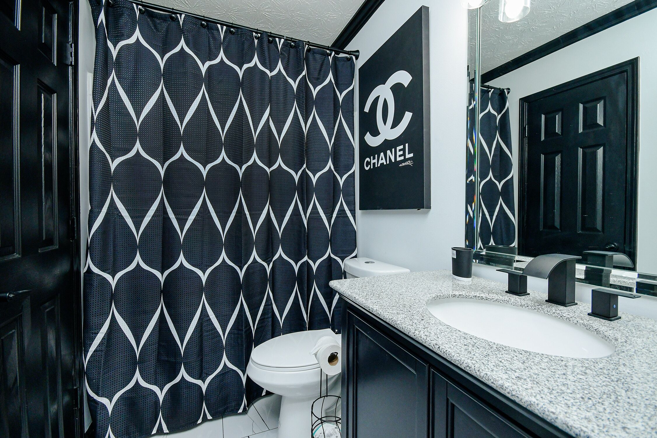 Ein Badezimmer mit einem schwarz-weiß gemusterten Duschvorhang, einer weißen Toilette, einem Waschbecken mit Granitplatte und einem Spiegel darüber.
