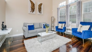 Ein modernes Wohnzimmer mit grauem Sofa, einem weißen Couchtisch und blauen Sessel. Es gibt zwei große Fenster mit Vorhängen, einen weißen Teppich und eine hohe Stehlampe.
