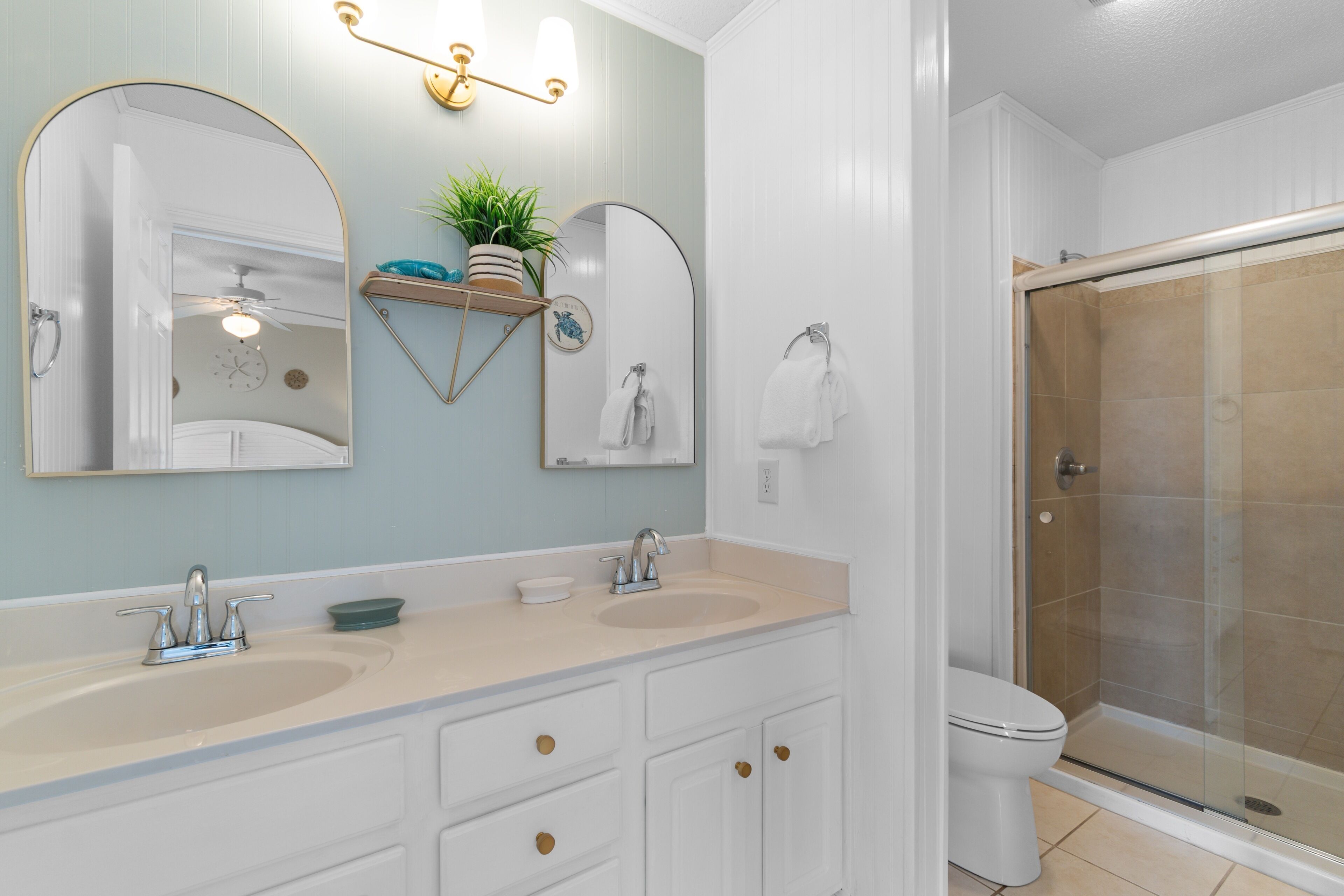 A bathroom with two sinks, a toilet, a shower, and a mirror with a plant on the wall.