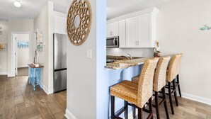 A modern kitchen with white cabinets, a stainless steel refrigerator, and a built-in microwave. There are three woven bar stools and a decorative wall piece. - Walk to Surfside Beach from This 4-Bedroom Condo w/ Resort Pool (Surfside Beach)
