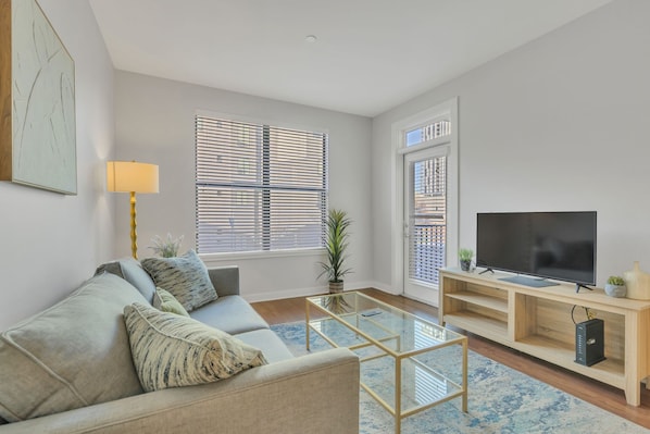 A modern living room with a sofa, a glass coffee table, a TV on a wooden stand, and a window with blinds.