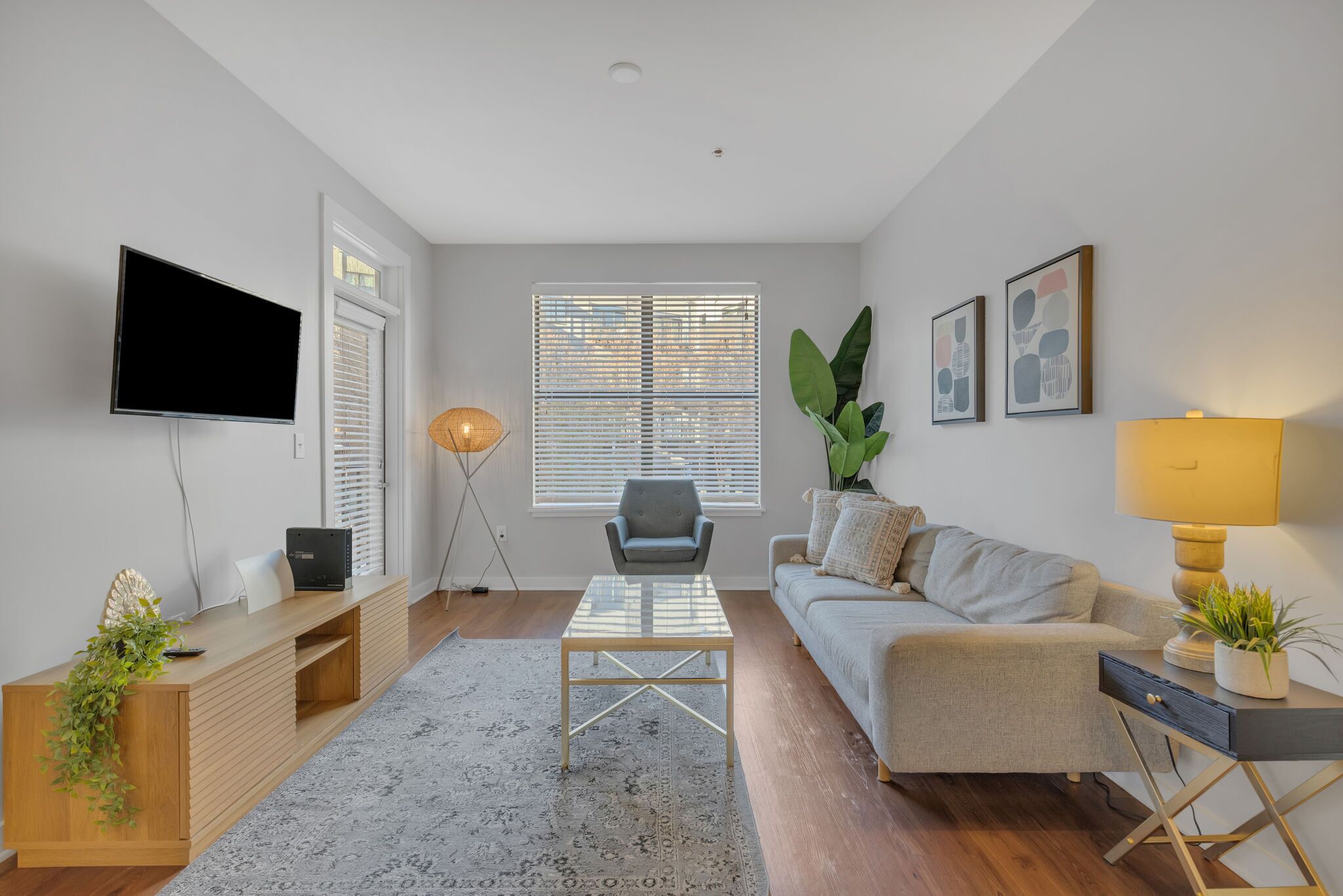 A modern living room with a flat-screen TV, a grey sofa, a coffee table, and a large window with blinds.