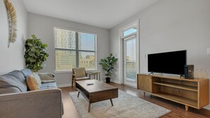Ein modernes Wohnzimmer mit grauem Sofa, Holztisch und Flachbildfernseher auf einem Holzständer. Zwei Fenster mit Jalousien, ein Blumentopf und ein kleiner Tisch mit Stuhl.