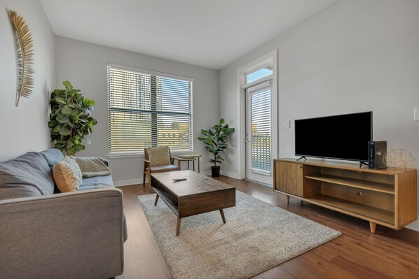 Una sala moderna con un sofá gris, una mesa de centro de madera y un televisor de pantalla plana sobre un mueble de madera. Hay dos ventanas con persianas, una planta en maceta y una pequeña mesa con una silla.