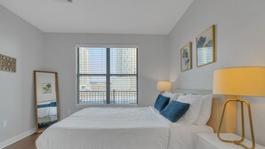 A bedroom with a bed, bedside table, lamp, and window with blinds. - Gulch Apartment with Gym 2BR/2BA (Nashville)