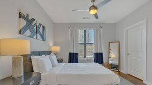 A modern bedroom with a bed, bedside lamps, a ceiling fan, a window with curtains, and a mirror.