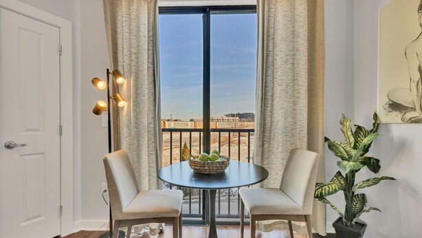 A room with a large window, two chairs, a table with a fruit bowl, a potted plant, and a painting on the wall.