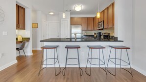 A modern kitchen with wooden cabinets, a stainless steel refrigerator, and a black countertop. There are three bar stools and a chair. - Perfect Pad in the Gulch! 2BR & Gym! (Nashville)