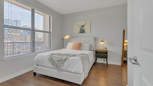 A bedroom with a large bed, two bedside lamps, a nightstand, a mirror, and a cityscape view through the window blinds. - Perfect Pad in the Gulch! 2BR & Gym! (Nashville)