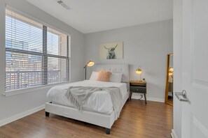A bedroom with a large bed, two bedside lamps, a nightstand, a mirror, and a cityscape view through the window blinds. - Perfect Pad in the Gulch! 2BR & Gym! (Nashville)