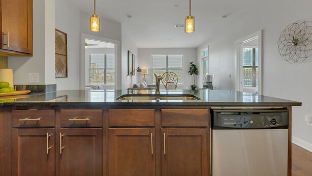 A modern kitchen with wooden cabinets, a stainless steel dishwasher, pendant lights, and a large island with a sink. - Perfect Pad in the Gulch! 2BR & Gym! (Nashville)