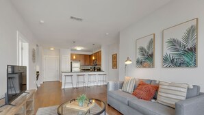 A modern living room with a grey sofa, a coffee table with a plant, and a TV on a wooden stand. A kitchen is visible in the background with wooden cabinets and a white countertop. - Perfect Pad in the Gulch! 2BR & Gym! (Nashville)