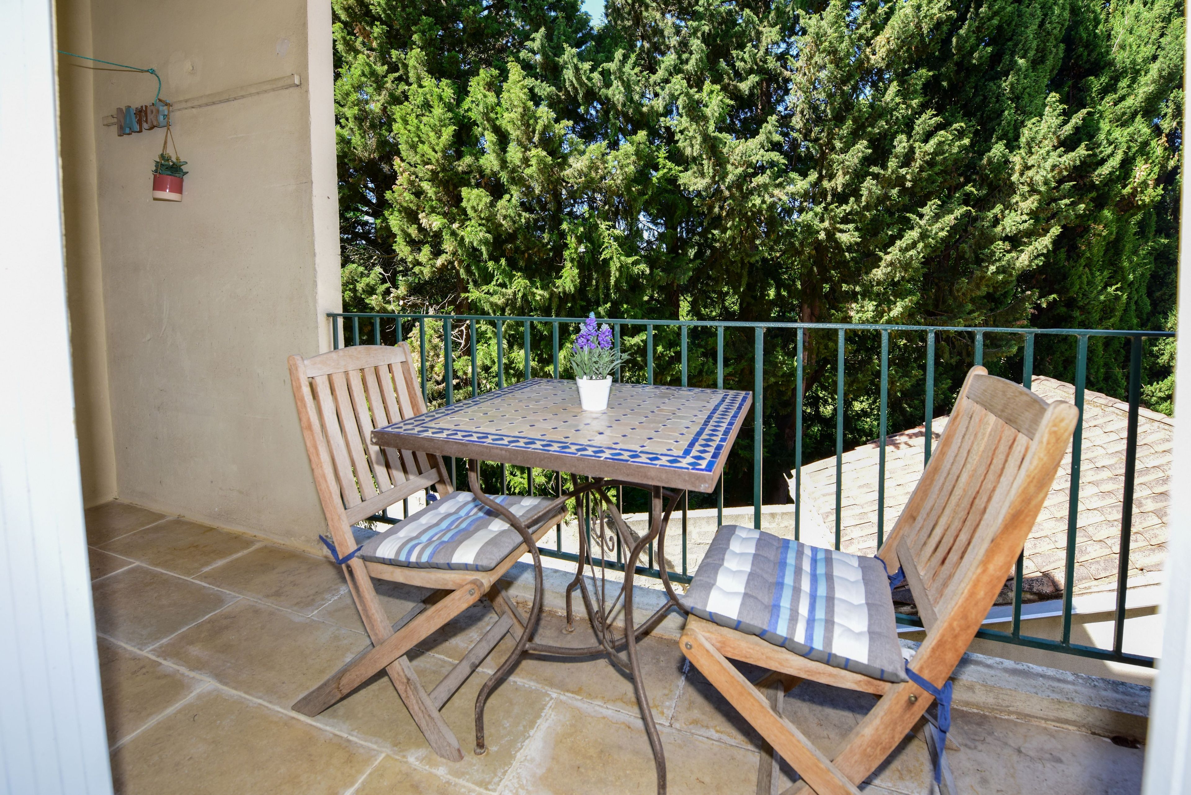 Ein Balkon mit Tisch und zwei Stühlen, umgeben von einem Metallgeländer und Grünpflanzen.