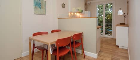 Un comedor con mesa de madera y sillas rojas, una cocina con un mueble blanco, y vistas al balcón exterior a través de una puerta de cristal.