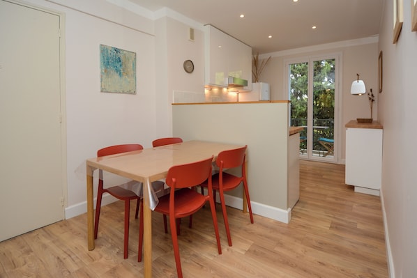 A dining area with a wooden table and red chairs, a kitchen area with a white cabinet, and a view through a glass door to an outdoor balcony. - Charming Renovated Apartment Near the Historic Center of Tarascon (Tarascon)