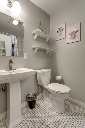 Une salle de bain avec un lavabo blanc, des toilettes et une poubelle. On y trouve deux étagères fixées au mur, ornées d’objets décoratifs, et un miroir au-dessus du lavabo.