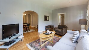 A modern living room with a flat-screen TV, a sofa, a coffee table, and a dining area with chairs. - Charming Hillsboro Home Near Vanderbilt (Nashville)