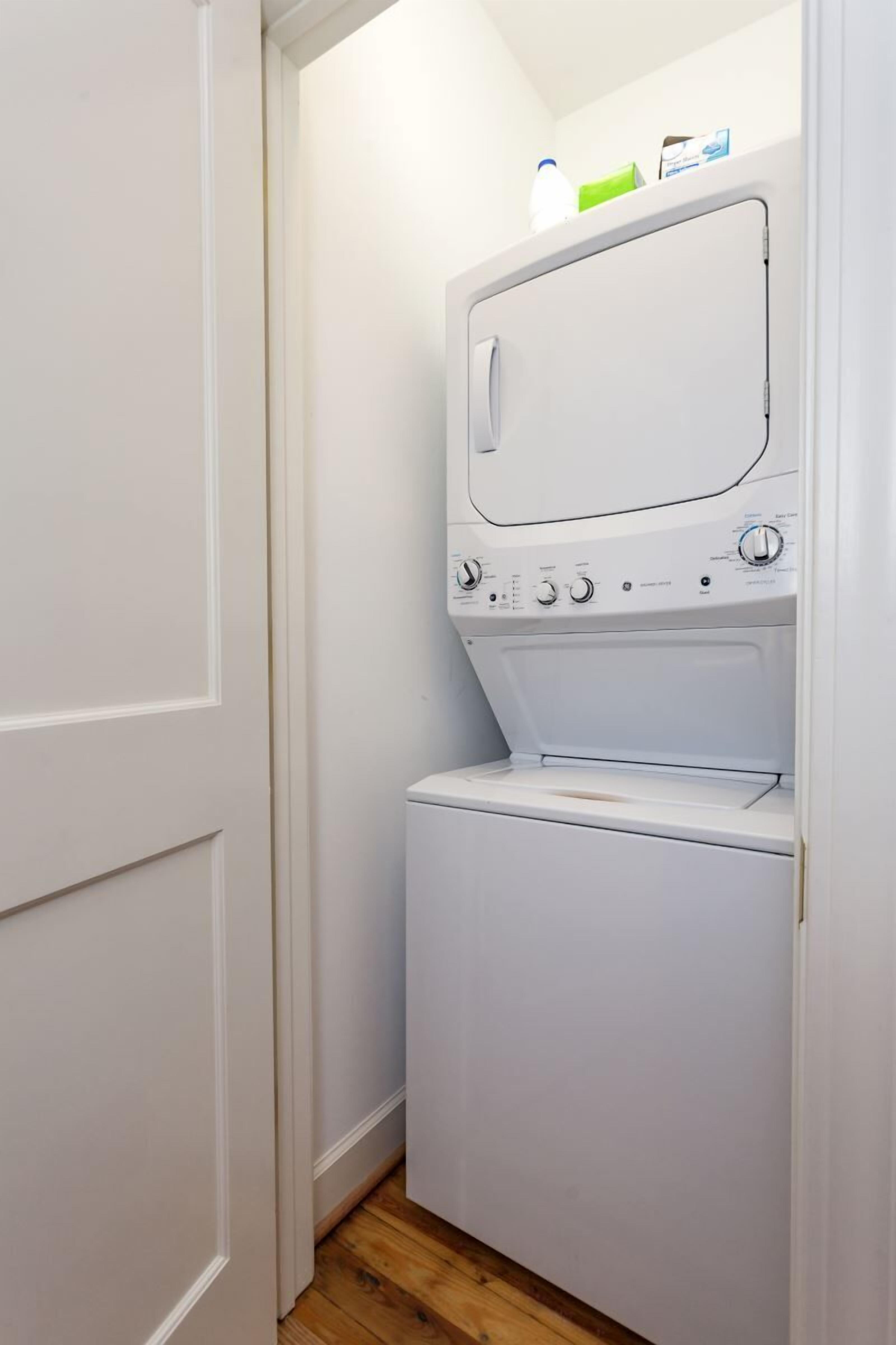 Une petite buanderie équipée d’un lave-linge et d’un sèche-linge à chargement frontal, d’une petite étagère avec une bouteille et une boîte, et d’un plancher en bois.