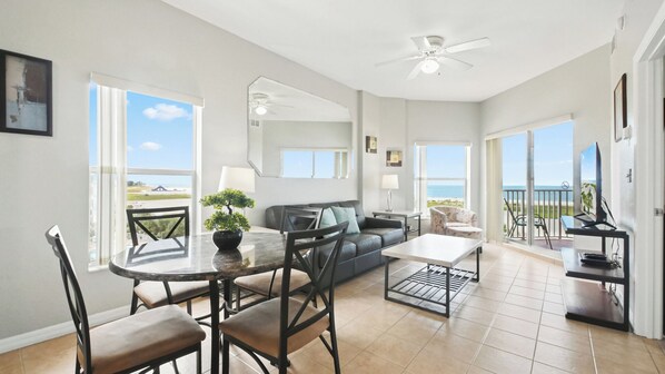 A spacious living room with a dining area, a sofa, and a TV. There are large windows offering a view of the beach.