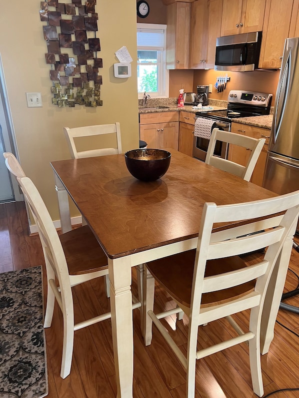 A dining area with a wooden table and white chairs, a decorative wall piece, and a kitchen in the background.