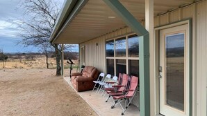 A covered porch with a brown leather chair and several folding chairs, a door with a screen, and a view of a dry landscape. - Field House on Lake Godstone (Graford)