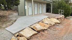 A portable restroom with four doors, a covered waiting area, and a concrete ramp leading up to it. - Field House on Lake Godstone (Graford)