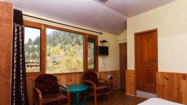 A room with wooden walls and a large window overlooking a forest. There is a bed, two chairs, and a small table. - Snow view Home Stay (Naggar)