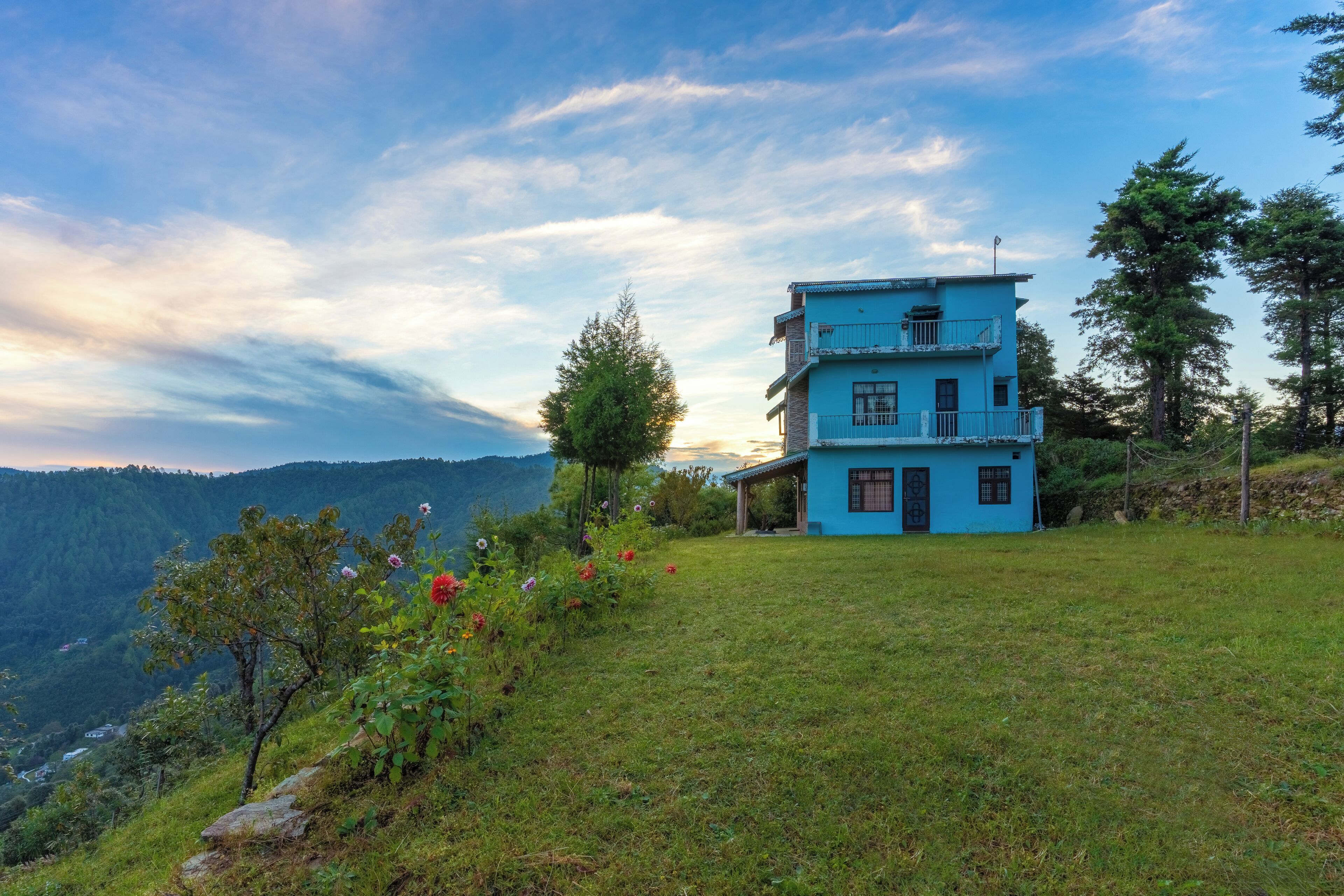 A two-story blue house with a balcony, surrounded by a grassy hill and trees, overlooking a valley.