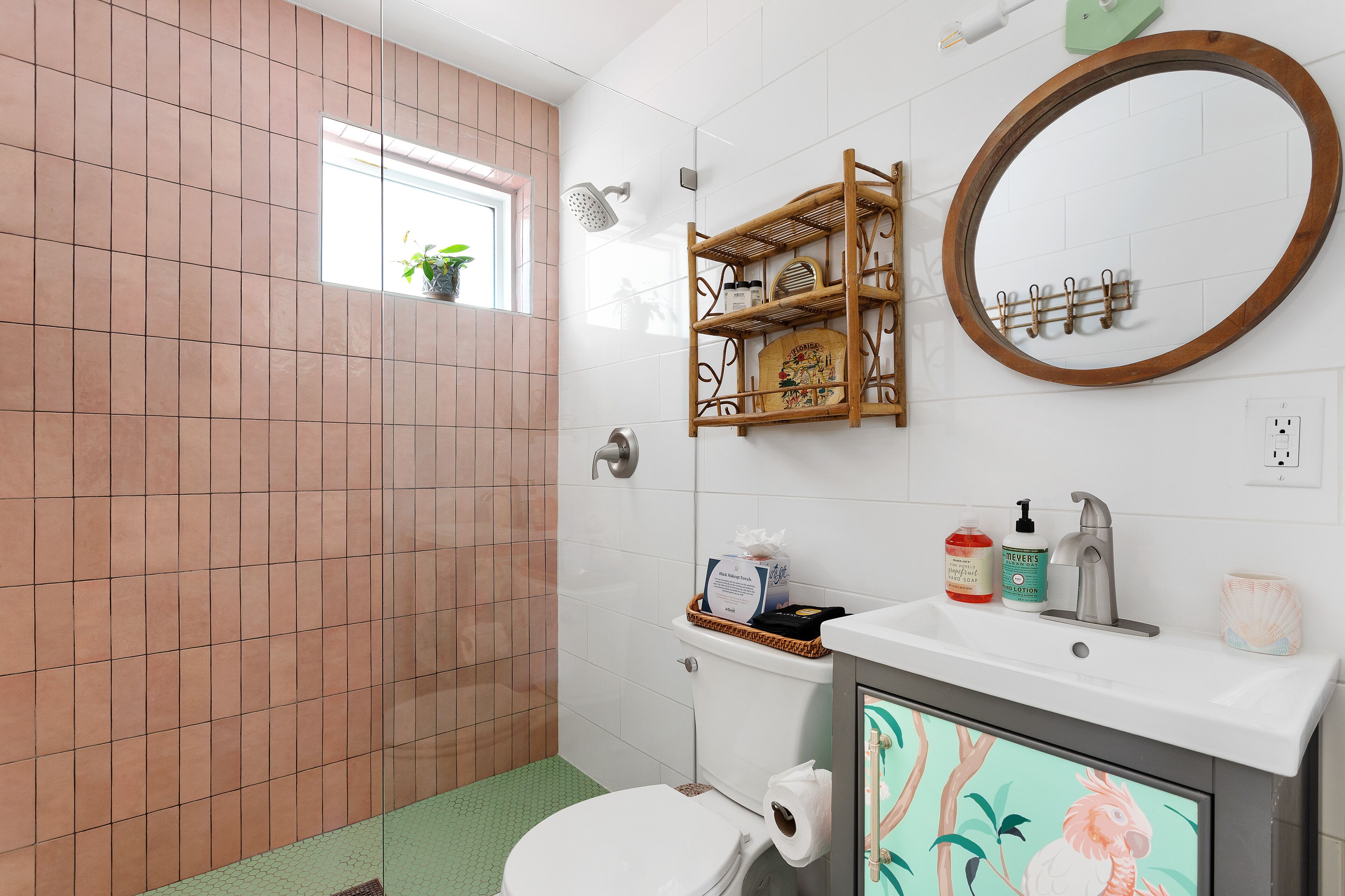 Een badkamer met toilet, wastafel en douche. Er is een raam met een plant, een plank met toiletartikelen en een spiegel.