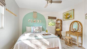 Un dormitorio con cama, cómoda, espejo, ventilador de techo y ventana con vista a árboles.