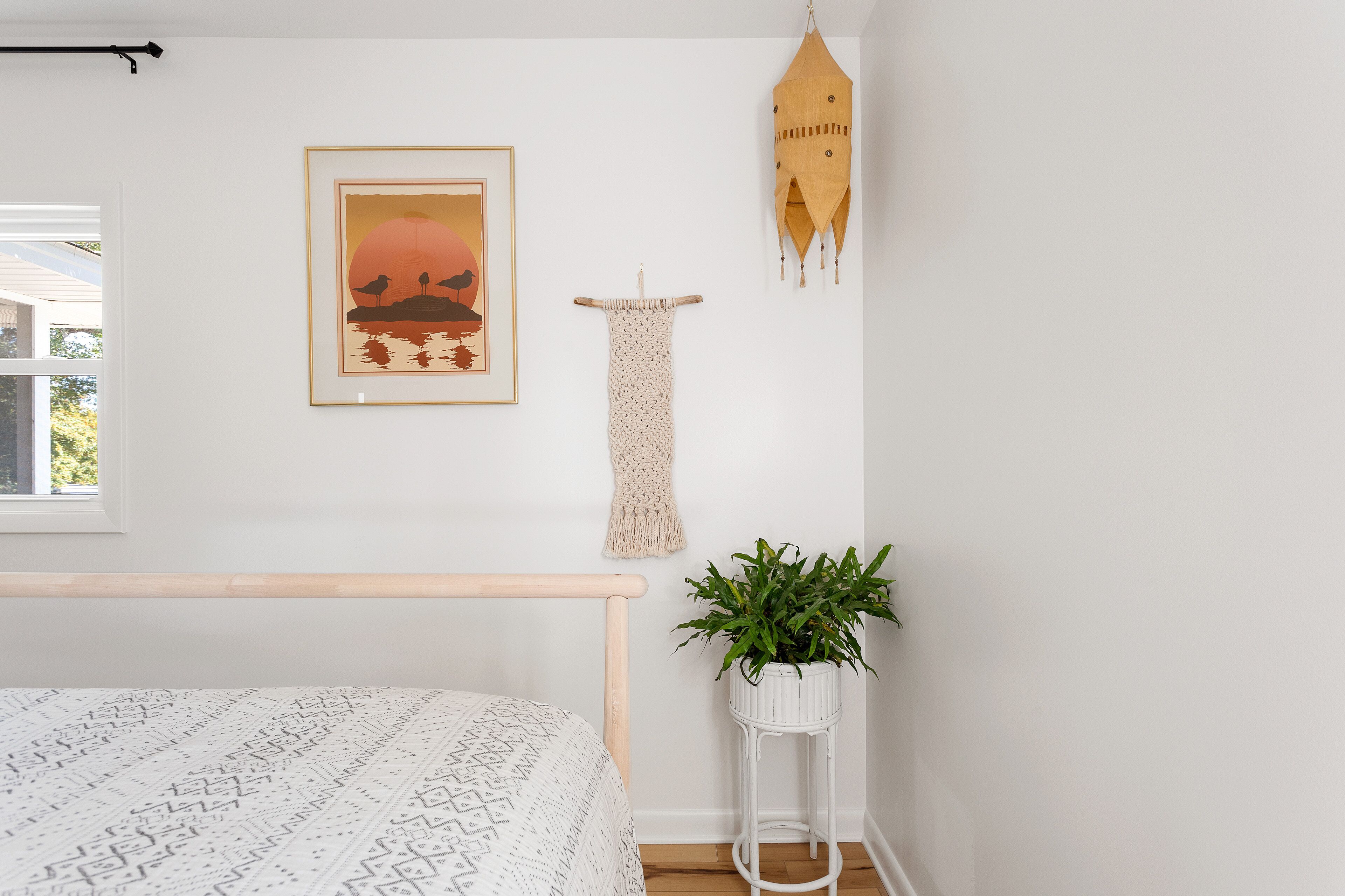 A bedroom with a bed, a wall-mounted macrame piece, a framed picture, a hanging macrame, a hanging macrame, and a potted plant.