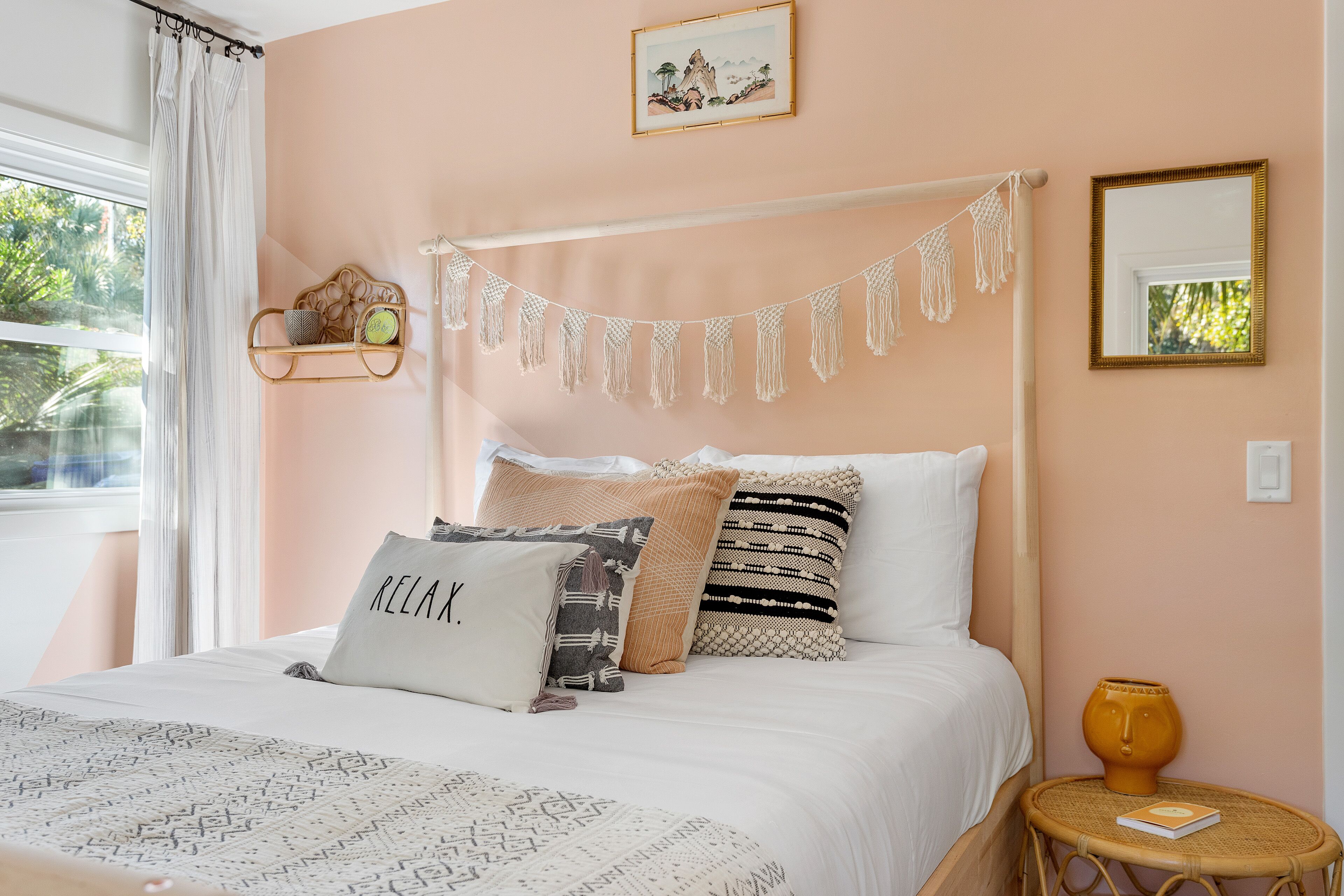 Un dormitorio con cama, mesita de noche, ventana con cortinas, un cuadro en la pared y un adorno de macramé.