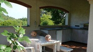 Une cuisine d’extérieur rustique, avec une table et un banc blancs, un petit réfrigérateur et une vue sur la verdure par une fenêtre.