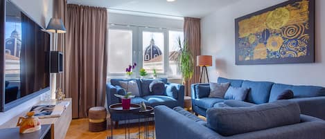 Ett modernt vardagsrum med en blå soffgrupp, en platt-TV och ett stort fönster med gardiner.