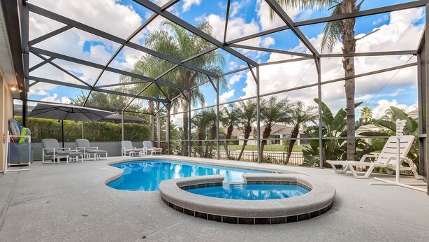 A covered outdoor swimming pool with lounge chairs and umbrellas, surrounded by palm trees and a clear sky. - Stunning Villa with Ultimate Games Room (Haines City)