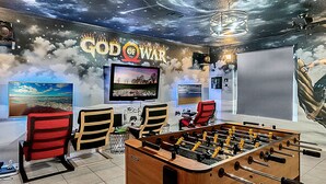 A room with a large screen, two gaming chairs, a foosball table, and a wall with a space-themed mural. - Stunning Villa with Ultimate Games Room (Haines City)
