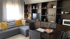 A modern living room with a grey sofa, yellow cushions, a dining table with chairs, a TV, and built-in shelves with various decorative items. - navas suite (Navas del Rey)