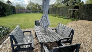 Un espace extérieur aménagé avec des sièges en bois, une table et des bancs, entouré d’une haie et d’une clôture en bois.